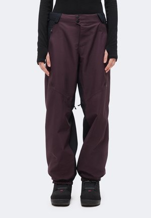 Personne portant un pantalon d’extérieur ample violet foncé avec des panneaux latéraux noirs, des bottes d’hiver noires et un haut noir ajusté à manches longues.