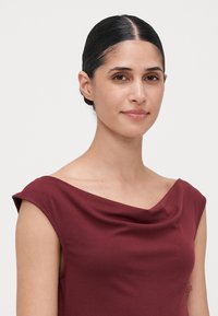 Mujer con pelo oscuro recogido hacia atrás, vistiendo una blusa sin mangas de color burdeos con un escote drapeado, mirando ligeramente sonriente hacia la cámara.