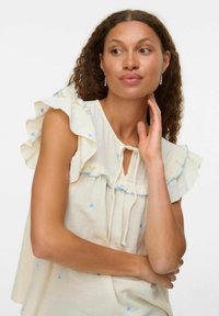 Blusa bianca a maniche corte con motivi a forma di cuore blu e spalline arricciate. Presenta un'apertura a chiave e un cordoncino al collo.