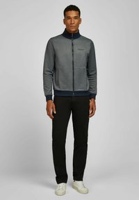 Man in een grijze zip-up jas, zwarte broek en witte sneakers staat tegen een effen lichtgrijze achtergrond.