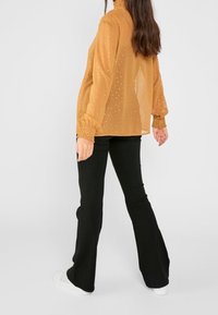 Blusa a maniche lunghe, trasparente, di colore senape con pois testurizzati, abbinata a pantaloni neri svasati e sneakers bianche.