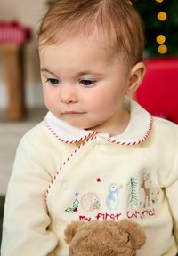 Vêtement couleur crème avec des bordures rouges, orné de broderies animales et de l'inscription "mon premier Noël". Texture douce ; enfant tenant un jouet en peluche.