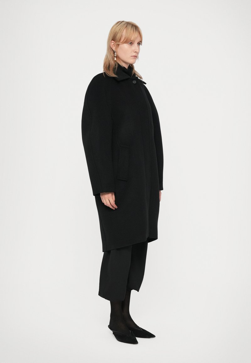 Svart ullkappa, överdimensionerad passform, hög krage, enkel knäppning, sidofickor, mjuk textur. Buren över svarta byxor och skor.