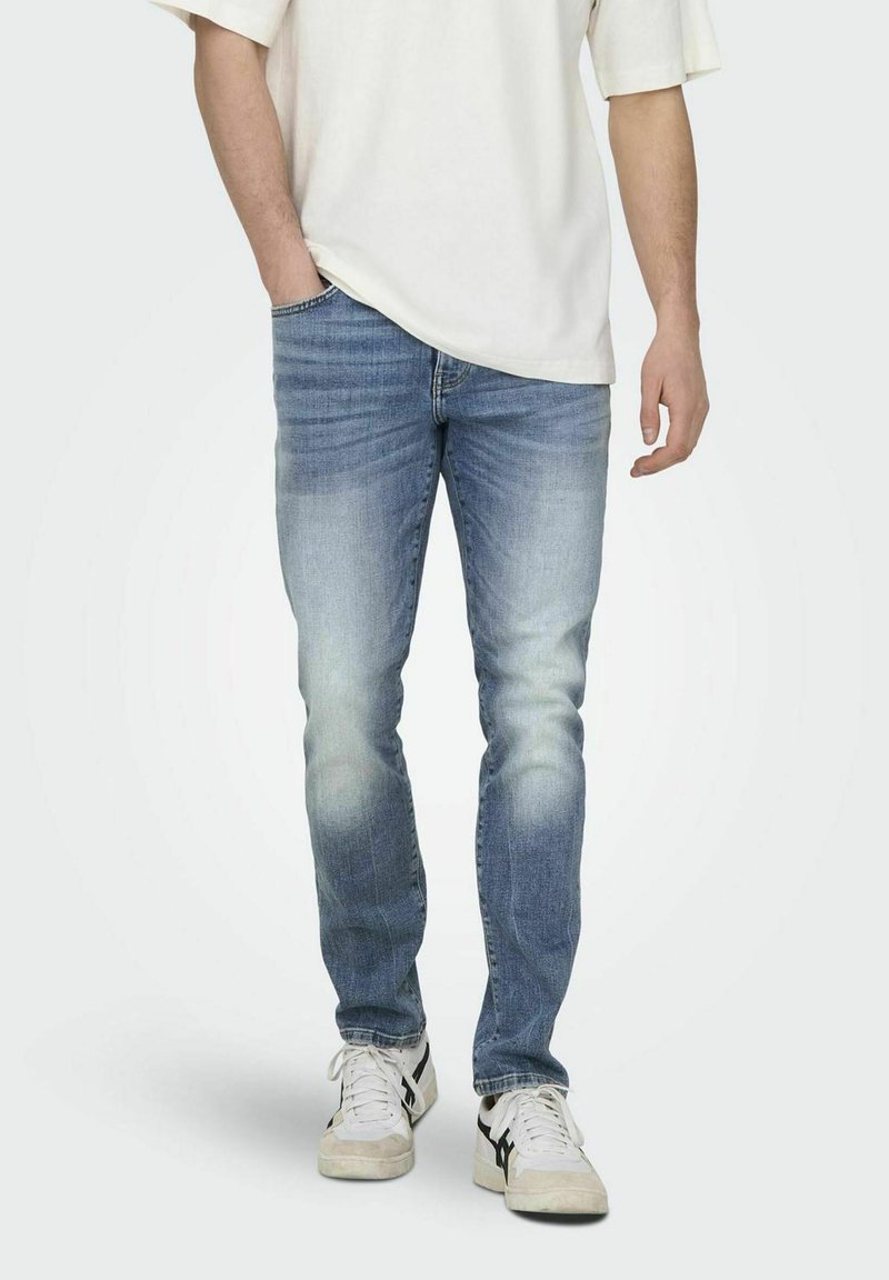 Lichtblauwe denim jeans in slim fit, vervaagd aan de voorkant, met vijf zakken. Gecombineerd met witte sneakers en een off-white shirt.