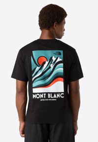 Hombre vistiendo una camiseta negra con un gr�áfico colorido de la montaña Mont Blanc, la silueta de un escalador, un sol rojo y el texto "Nunca Dejes de Explorar" en la parte de atrás.