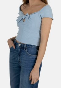 Donna che indossa un top a spalle scoperte azzurro chiaro a coste con dettaglio a fiocco frontale e jeans in denim blu a vita alta, con una mano in tasca.