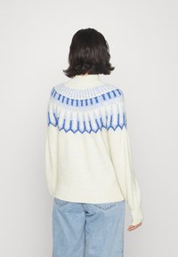 Person with dark hair wearing a white sweater with blue patterned yoke and light blue jeans, facing away against a plain white background.