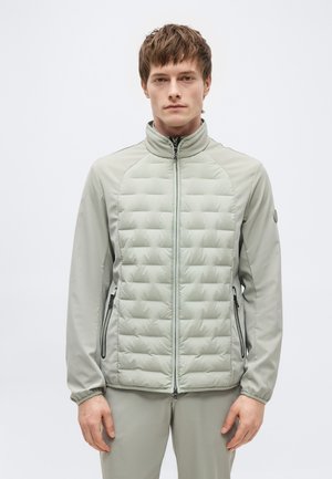 Νέος άνδρας φοράει ανοιχτό γκρι μπουφάν με φερμουάρ σε καπιτονέ ύφασμα και ταιριαστό παντελόνι, στέκεται μπροστά σε απλό λευκό φόντο.