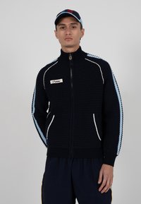 Veste bleu marine zippée avec tricot texturé, rayures bleu clair et blanches le long des manches. Comprend des poches latérales et un patch logo sur la poitrine.