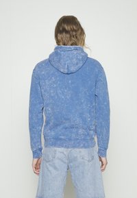 Sudadera azul hecha de suave algodón. Presenta un corte holgado, capucha con cordones y una textura deslavada. La vista desde atrás muestra puños y dobladillo acanalados.