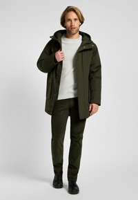 Pikowana kurtka parka w ciemnozielonym kolorze z kapturem, zapinana na zamek z przodu i z bocznymi kieszeniami, nałożona na jasnoszary sweter i ciemnozielone spodnie.