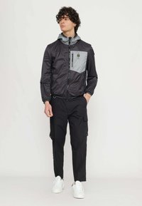 Giacca leggera nera con accenti grigi e cappuccio, dotata di una tasca con zip. Abbinata a pantaloni cargo neri e sneakers bianche.