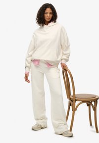 Cremefarvet oversized hættetrøje med forlomme, kombineret med matchende bukser med vide ben. Tekstureret materiale med en lag-på-lag lyserød stribet skjorte under.