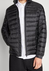 Osoba v černé prošívané zapínací bundě přes bílou košili s rukama v kapsách, zobrazená od krku po boky.