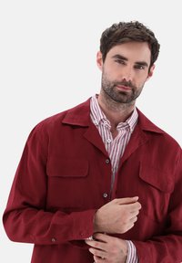 Rotes Leinenhemd-Jackett mit zwei Brusttaschen, Knopfleiste und leichtem Stoff. Darunter ist ein weißes und rotes gestreiftes Hemd sichtbar.