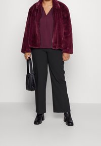 Veste en fausse fourrure bourgogne avec une fermeture éclair, associée à un haut sombre et un pantalon noir à jambes larges. Bottes noires à talons épais et un petit sac.