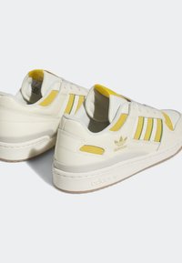 Zapatillas de cuero blancas con acentos en amarillo y verde, que cuentan con tres franjas, una suela texturizada y un detalle de logo en el lateral exterior.
