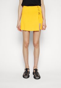 Mini-jupe jaune côtelée avec un détail de boucle latérale, complétée par des sandales noires à brides avec des accents métalliques et une semelle texturée.