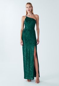 Groene pailletten jurk met een asymmetrische halslijn, zijspleet en aansluitend ontwerp. Draagt een huidkleurige schoen, tegen een lichte achtergrond.