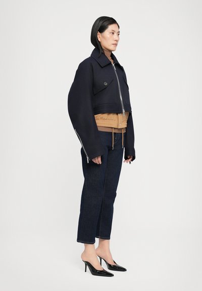 Chaqueta de marinero recortada con mangas anchas, cremallera plateada y bolsillos frontales sobre una capa de plumas color beige, combinada con jeans oscuros y tacones negros.