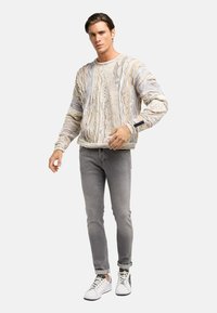 Textuurtrui in lichte kleur met golvende patronen, gedragen met grijze jeans en witte sneakers met zwarte accenten.
