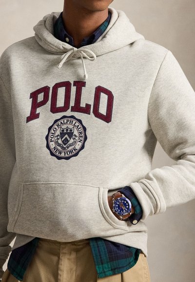 Άτομο που φοράει ανοιχτό γκρι φούτερ Polo Ralph Lauren πάνω από ένα μπλε-πράσινο καρό πουκάμισο, μπεζ παντελόνι και ένα ρολόι με μπλε καντράν και καφέ λουράκι.
