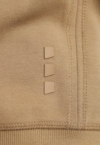 Tessuto a maglia beige con tre piccole toppe quadrate allineate verticalmente, con cuciture visibili lungo la cucitura.