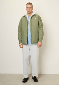 Lys grøn quiltet jakke med grå hætteforing, båret over en blå skjorte og grå sweatpants. Sorte sko fuldender outfittet.