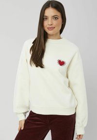 Sweatshirt léger crème avec un col rond et des manches longues, froncées ; arbore une broderie en forme de cœur rouge sur la poitrine. Associé à un pantalon en velours côtelé rouge foncé.