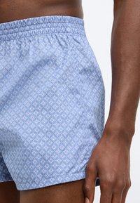 Ανοιχτό μπλε σορτς με ολόκληρο λουλουδάτο σχέδιο, διαθέτει ελαστική μέση και απαλή υφή. Κοντινή λήψη που δείχνει λεπτομέρεια και εφαρμογή.