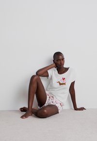 Camiseta blanca con un gráfico de teckel y corazones, combinada con pantalones cortos estampados que presentan corazones en rojo y rosa. Tela de algodón, ajuste casual.