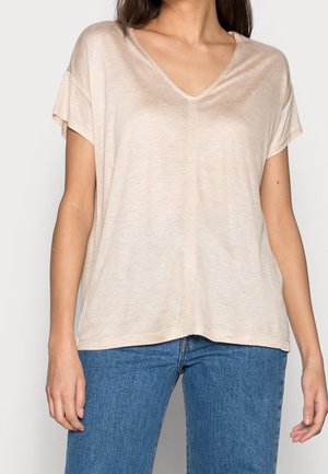 Kvinde iført beige V-hals T-shirt med korte ærmer og løs pasform, kombineret med blå jeans, foran en ensartet lys baggrund.