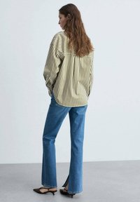Camicia oversize a righe gialle e marroni, jeans flared blu e tacchi marroni. I jeans hanno delle fessure laterali. Capelli lisci e strutturati che cadono sulla schiena.