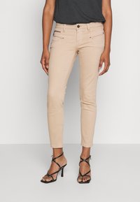 Jeans skinny beiges clairs à taille mi-haute, avec des poches avant et des accents métalliques. Associés à des sandales à talons aiguilles noires à lanières.