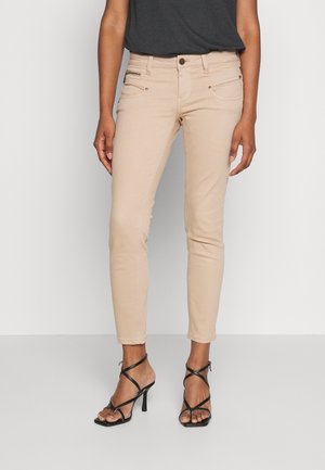 Jeans skinny de color beige claro con cintura de media altura, que presentan bolsillos frontales y detalles en metal. Combinados con sandalias de tacón alto negras con correas.
