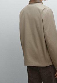 Lichtbruin jasje met een gestructureerd ontwerp, voorzien van een bruine kraag en een gladde textuur. Voorzien van zijsplitten voor extra comfort.