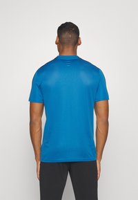Camiseta atlética azul de manga corta con una textura suave y cuello redondo. Presenta detalles negros en los hombros. Tejido ligero.