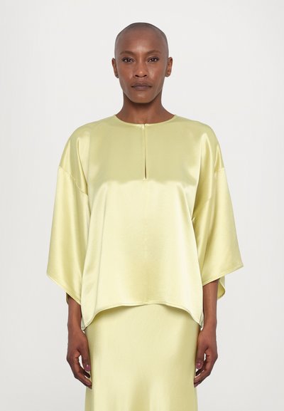 Blouse satin jaune avec un décolleté bateau, épaules tombantes et une petite fente à l'avant. Design fluide, se marie avec une jupe assortie.