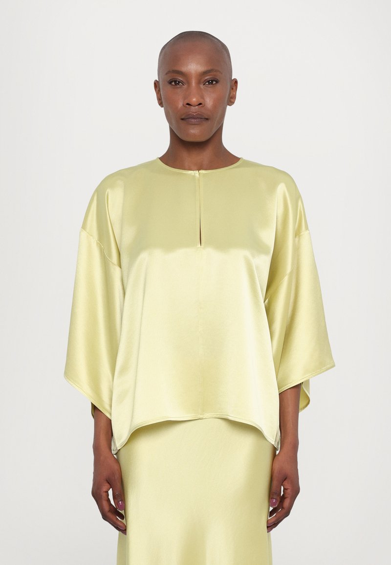Blusa amarilla de satén con escote barco, mangas caídas y una pequeña abertura en el frente. Diseño fluido, combina con una falda a juego.