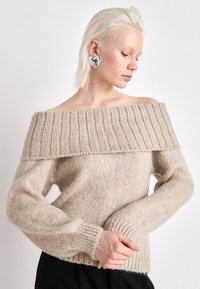 Beige stickad tröja med off-shoulder design, ribbad halsringning, långa ärmar och textilstruktur; matchad med en silverhjärtörhänge.