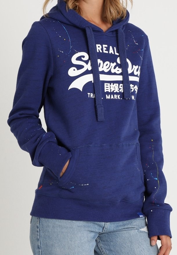 Personne portant un sweat à capuche bleu Superdry avec un logo blanc et des détails en peinture éclaboussée, associé à un jean bleu clair déchiré.