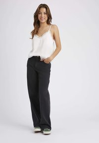 Camisole blanche en satin avec des dentelles, associé à un jean noir taille haute à jambes larges et des baskets avec des accents verts. Le modèle se tient les mains dans les poches.