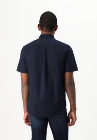 Camicia blu navy a maniche corte con colletto standard, piega posteriore per una vestibilità migliore e una texture liscia, abbinata a jeans in denim scuro.