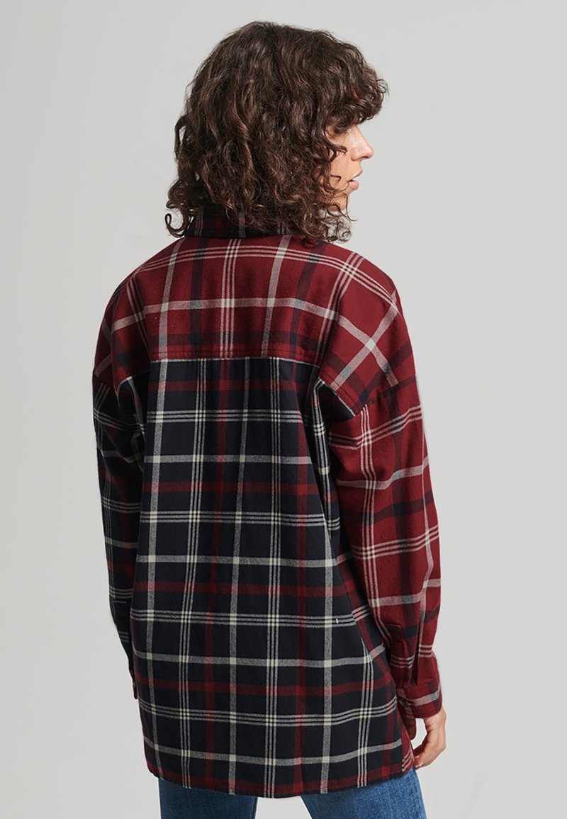 Personne aux cheveux bouclés portant une chemise à manches longues oversize à carreaux rouges et noirs, debout de profil sur un fond uni.
