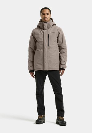Man draagt een taupekleurige winterjas met capuchon, zwarte broek en zwarte wandelschoenen, staand tegen een effen lichte achtergrond.