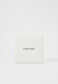 Baltas stačiakampis dežė su lygiu paviršiumi, ant priekio centruotas juodas serifinis šriftas su "Calvin Klein" logotipu.