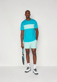 Camisola desportiva turquesa com uma listra azul claro, combinada com calções a condizer. O modelo segura uma raquete de ténis, usando meias brancas e sapatos azul-marinho.