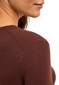 Nahaufnahme einer Personenschulter und des Nackens, die einen strukturierten dunkelbraunen Pullover trägt, vor einem schlichten weißen Hintergrund.