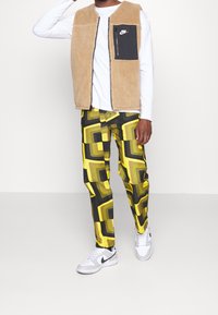 Beige fleeceställ över en vit långärmad skjorta, kombinerat med mönstrade gula och svarta byxor samt vita Nike-sneakers.