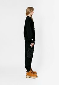 Sudadera negra de gran tamaño, pantalones joggers negros con bolsillo lateral, botas marrones con suelas gruesas. Cabello suelto, de perfil, contra un fondo blanco.